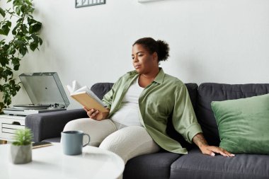 Afro-Amerikalı bir kadın, şık oturma odasında kitap okuyarak evde vakit geçirmekten zevk alıyor..