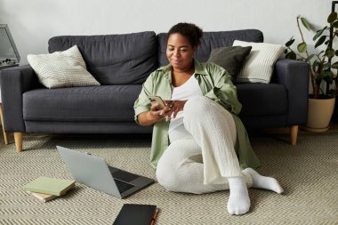 Dairesinde rahat bir şekilde çalışan bir kadın, yerde otururken telefonunu kontrol ediyor..