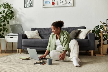 Afro-Amerikalı bir kadın şık bir dairede dizüstü bilgisayarını kullanarak yerde dinleniyor..