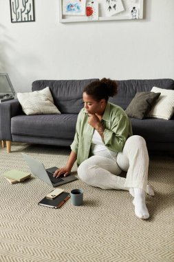 Sıradan giyinmiş Afrikalı Amerikalı bir kadın yerde otururken dizüstü bilgisayarına yoğunlaşıyor..