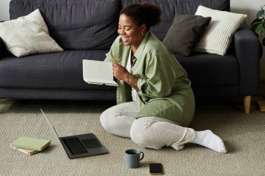 Afro-Amerikalı bir kadın yerde oturmuş, gülümseyip yazarken uzaktan kumandayla iş yapmaktan zevk alıyor..
