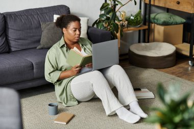 Afro-Amerikalı bir kadın evinde rahat bir şekilde otururken dizüstü bilgisayarı üzerinde özenle çalışıyor..