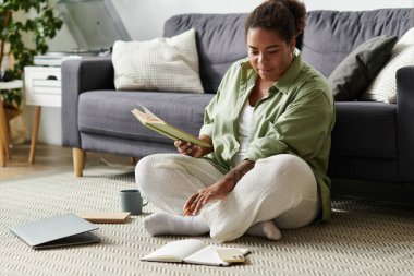 Gündelik giyinmiş genç bir kadın rahat dairesinde kitap okumaktan ve not almaktan hoşlanır..