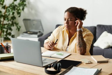 Bir kadın masasında oturmuş, rahat bir dairede dizüstü bilgisayarıyla düşünceli bir şekilde çalışıyor..