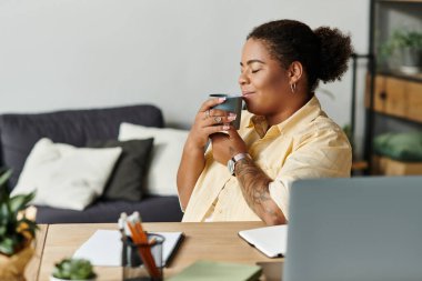 Afro-Amerikalı bir kadın rahat dairesinde uzaktan çalışırken kahvesini yudumluyor..