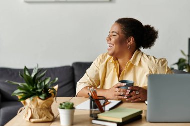 Rahat bir Afrikalı Amerikalı kadın şık bir dairede laptopuyla çalışırken kahvesini yudumluyor..