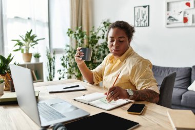 Afrikalı bir Amerikalı kadın, evinde bir kupa ve dizüstü bilgisayarı dengede tutarken notlar yazıyor..