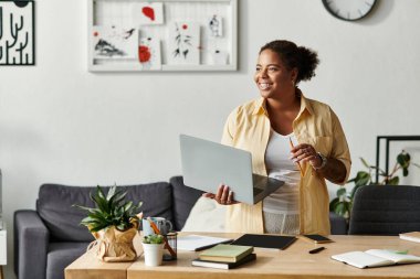 Afro-Amerikalı bir kadın laptopuyla şık bir ofis ortamında çalışırken gülümsüyor.