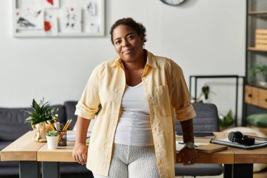 Afro-Amerikalı bir kadın rahat bir şekilde evde çalışır, spor kıyafetleri giyer ve kendine güvenen bir gülümsemesi vardır..