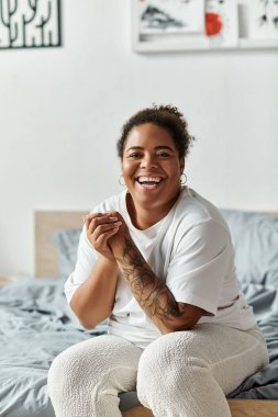 Afro-Amerikalı bir kadın sıcak bir dairede yatağında oturmuş gülümsüyor ve sabahının tadını çıkarıyor..
