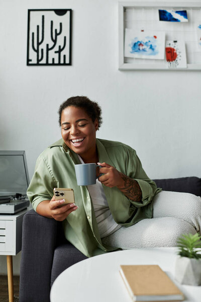 Афроамериканка наслаждается своей уютной квартирой, потягивая кофе, глядя в телефон.