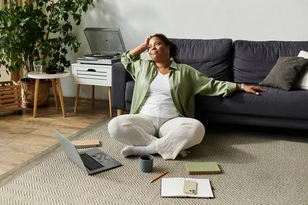 Sıradan giysiler içinde Afrikalı Amerikalı bir kadın yerde oturup uzaktan çalışma ortamının tadını çıkarıyor..