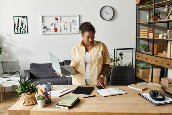 An African American woman in casual clothes multitasks on her laptop in a modern apartment.