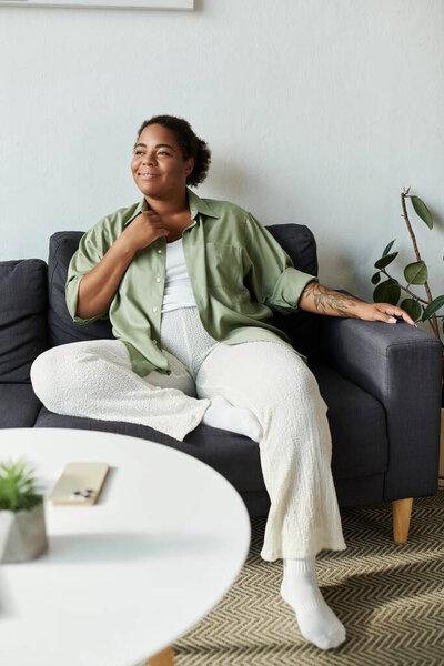 Афроамериканка в повседневной одежде счастливо сидит в своей стильной квартире.
