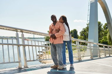 Neşeli genç çift, açık mavi gökyüzünün altında suyun kenarında gezinirken, sevgi dolu bir anı paylaşıyor..