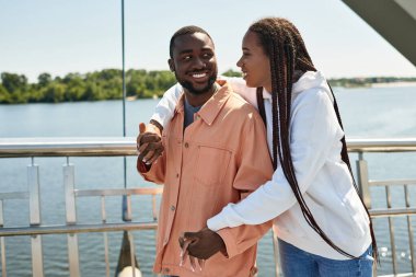 Genç bir çift su kenarında el ele yürümekten ve neşeyle gülümsemekten zevk alır..