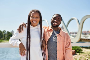 Neşeli bir çift, güneşli gökyüzünün altında, nehrin kıyısında el ele yürüyor..