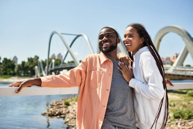 Erkek ve kadın, güneşli bir günde nehir kenarında yürürken sıcacık gülümserler..