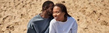 Genç bir çift, güneşli bir kumsalda el ele gülüşmeler ve kahkahalar atarak yürüyor..