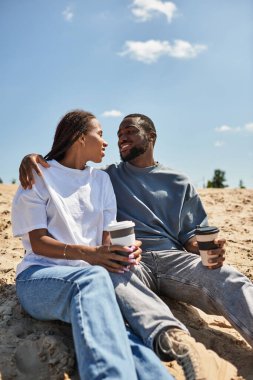 Bir çift açık havada mutlu bir şekilde yürür, açık mavi gökyüzünün altında gülücükler ve içkileri paylaşır..