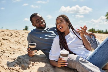 Genç bir çift, mutlu bir şekilde kumların üzerinde rahatlıyor, kahvelerini yudumluyor ve keyifli anlarını paylaşıyorlar..