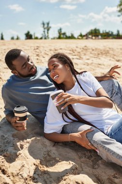 Sevgi dolu çift kumlu sahilde rahatlar, kahvelerini yudumlar ve eğlenerek birbirlerine eşlik ederler..