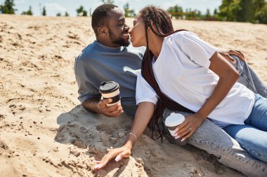 Genç çift sıcak kumda dinlenirken, kahvelerini yudumlarken ve gülümserken tatlı bir öpücüğü paylaşıyor..