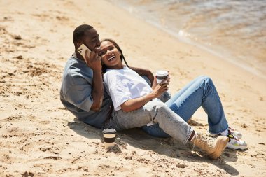 Genç çift kumsalda dinleniyor, sıcak içecekler içerken kahkahalarını ve sevgilerini paylaşıyor..