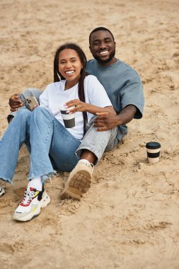 Sevgi dolu bir çift kumlu sahilde oturur, gülümsemelerini ve sıcak içkilerini paylaşarak eğlenirler..