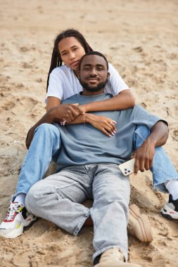 Genç çift kumsalda sıcacık sarılıyor, yumuşak kum ve renkli gökyüzü ile çevrili..