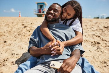 Genç çift sıcak kumda otururken, sevgi ve mutluluğu yakalarken neşeli bir kucaklaşmayı paylaşıyor..