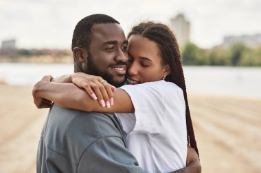 Genç bir çift su kenarında el ele dolaşır, gülümseyerek yüzlerini aydınlatır..