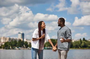Genç çift, güneşli bir günde suyun kenarında yürürken el ele tutuşup kahkahalarını paylaşıyor..