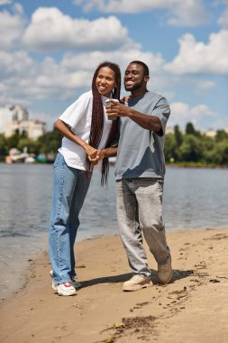 Neşeli çift nehir kıyısında yürüyor, açık mavi gökyüzü altında birbirlerine eşlik ederek eğleniyorlar..
