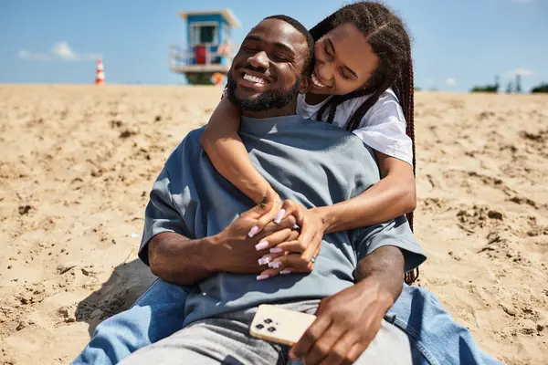 Genç çift sıcak kumda otururken, sevgi ve mutluluğu yakalarken neşeli bir kucaklaşmayı paylaşıyor..