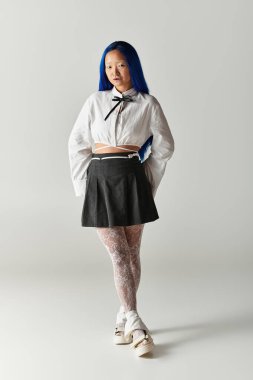 Mavi saçlı kendine güvenen genç bir Asyalı kadın gösterişli gri bir stüdyoda şık bir kıyafet sergiliyor..