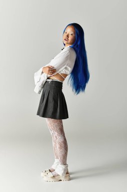 Modaya uygun, mavi saçlı genç bir kadın gri bir stüdyoda dikilirken şık elbisesini sergiliyor..