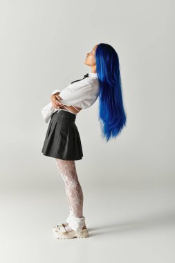 Gri bir stüdyoda zarif poz veren genç bir kadın şık elbisesi ve mavi saçlarıyla hava atıyor..