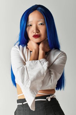 Güzel genç bir kadın, şık gri bir stüdyoda mavi saçlarını ve şık kıyafetlerini sergiliyor..