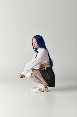 Canlı mavi saçlı genç bir kadın, gri stüdyo ortamında kendine güvenerek poz veriyor. Modayı somutlaştırıyor..