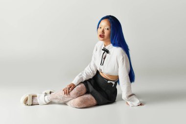 Mavi saçlı genç bir kadın modern gri bir stüdyoda zarif pozlar veriyor..