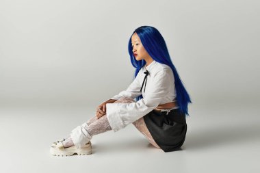 Gri bir stüdyoda, genç bir kadın çarpıcı mavi saçlarını ve şık kıyafetlerini gösteriyor..