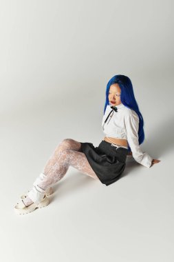 Şık genç bayan zarif bir şekilde oturuyor, eşsiz mavi saçlarını ve şık kıyafetlerini sergiliyor..