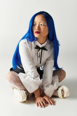 Gri bir stüdyoda genç bir kadın eşsiz mavi saçlarını ve şık kıyafetlerini gösteriyor..