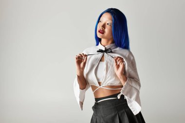 Şık genç bir kadın gri bir stüdyoda poz veriyor, çarpıcı mavi saçlarını ve moda kıyafetlerini sergiliyor..