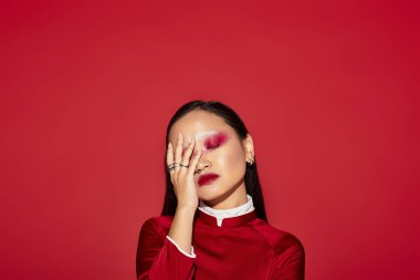 Genç Asyalı bir kadın kırmızı bir zemine karşı canlı göz makyajı ve modaya uygun kıyafetler sergiliyor.