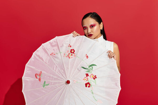 Молодая азиатка с ярким макияжем глаз уверенно позирует на ярком красном фоне.