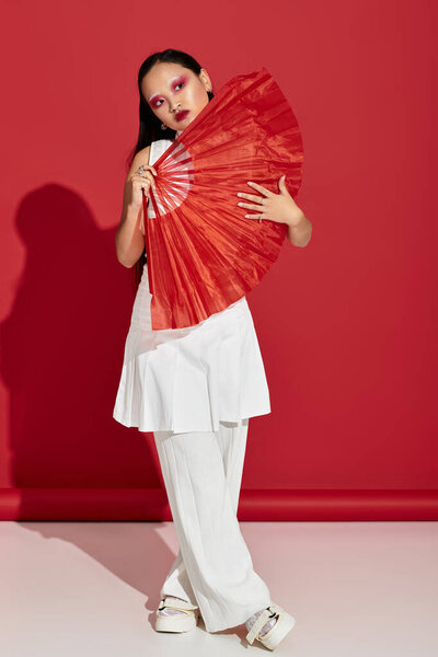 A young asian woman stands confidently in a red studio, displaying stylish fashion and artistic makeup.