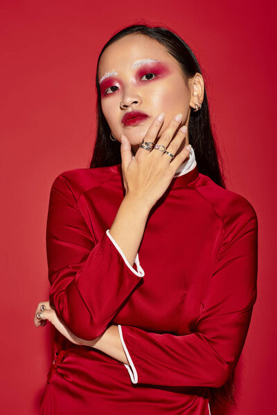 Молодая азиатка уверенно позирует в модных нарядах со смелым макияжем на красном фоне.