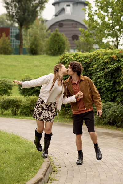 A happy young couple walks hand in hand, sharing a loving kiss in a vibrant park setting.